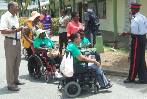 Two women in wheelchairs are on the road approaching a high curb, they are surrounded by friends and supporters