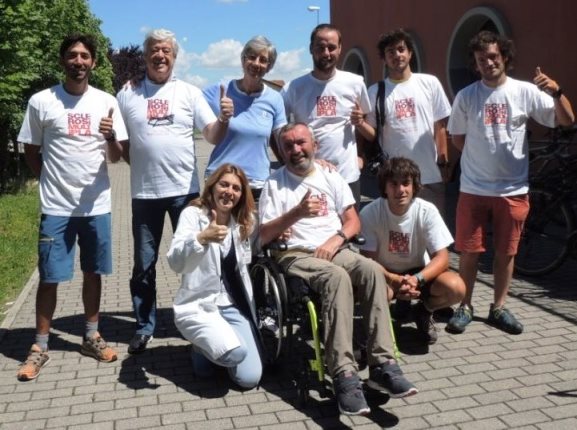 A group of people post for a photo. It's a sunny day and everyone is smiling and giving a thumbs up. One man is in a wheelchair and two people crouch down beside him.